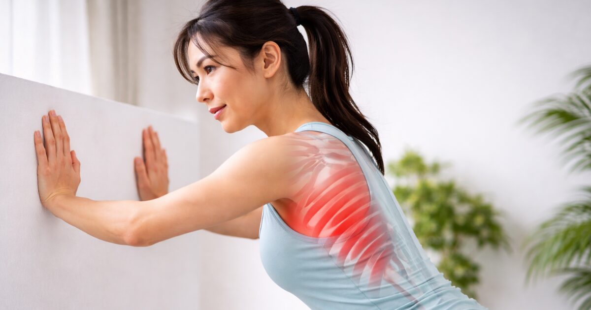 前鋸筋を意識して壁を押す女性の横姿。脇から肋骨外側の筋肉がハイライトされている