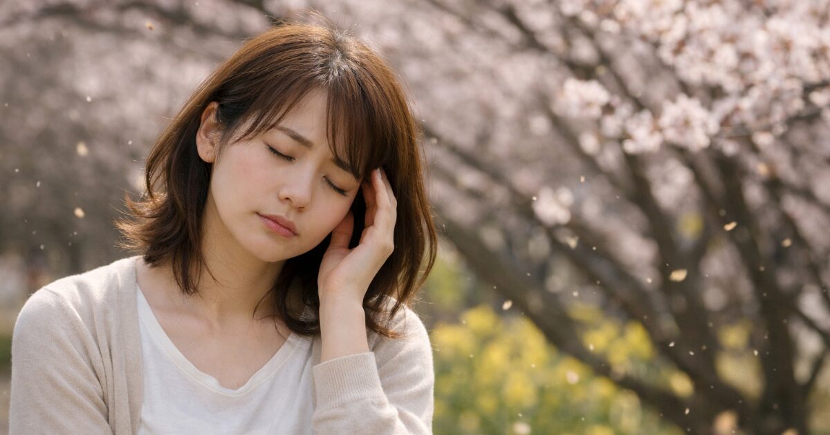 春の桜の下で頭に手を当て体のだるさを感じている女性のイメージ